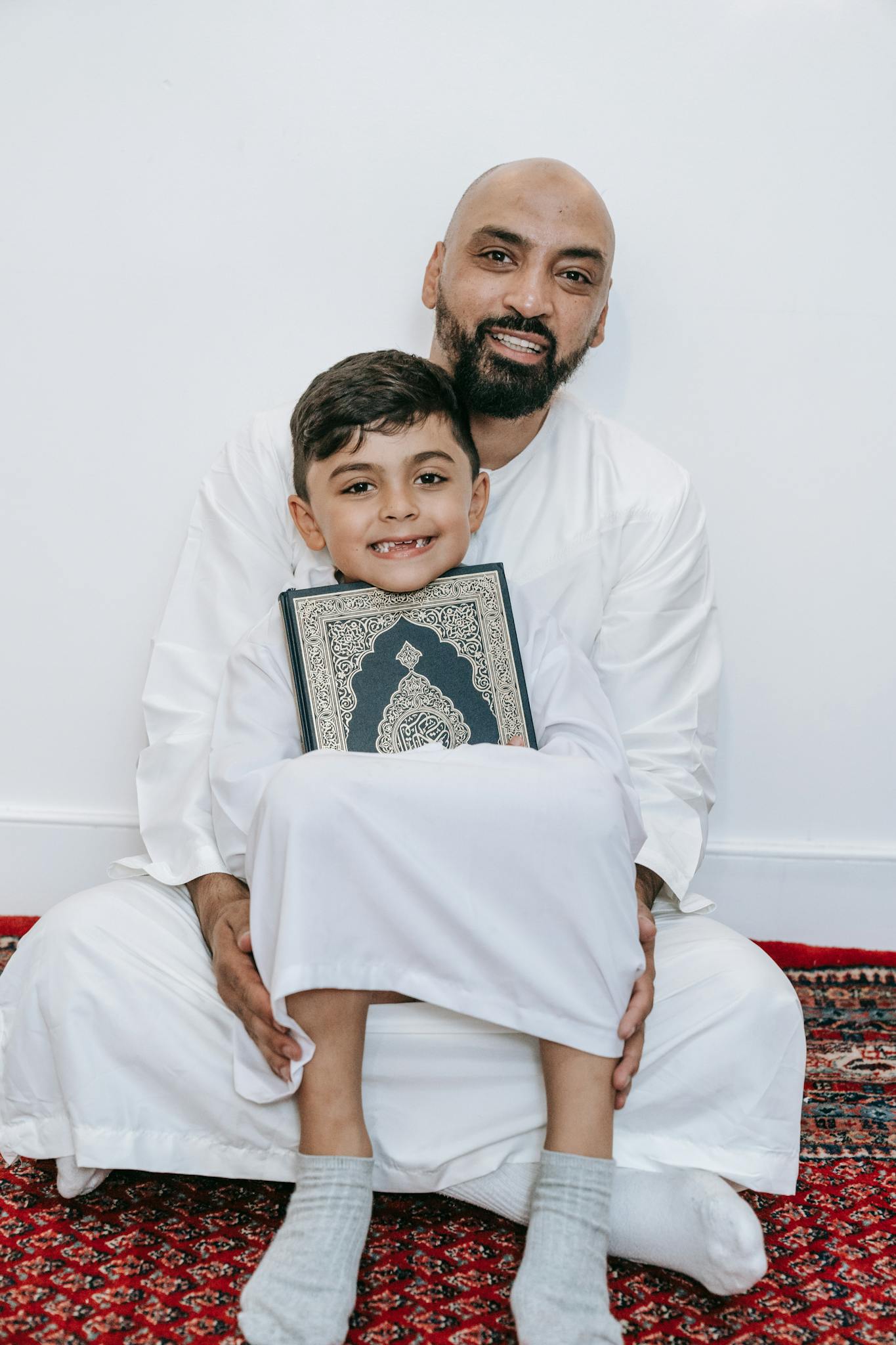 A father and son smiling, holding a holy book while wearing traditional attire.