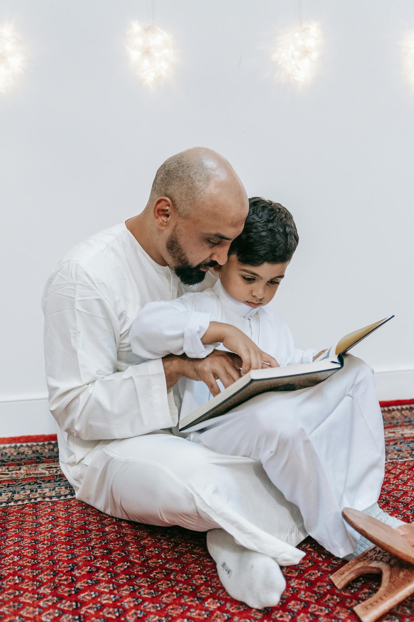 A father and son wearing traditional attire reading a book together inside a cozy room.