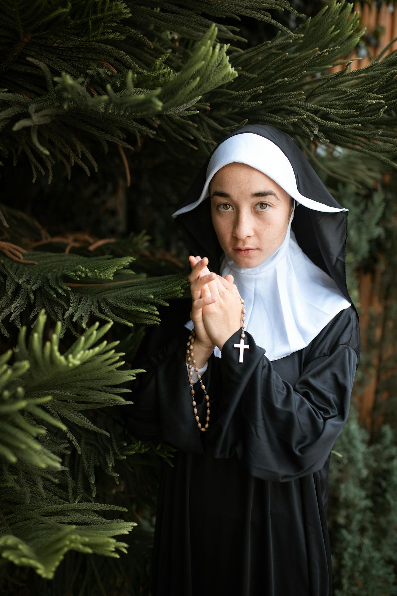A nun in traditional habit holds a rosary near a tree, symbolizing faith and devotion.