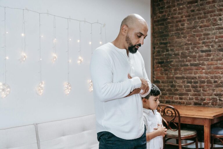 Father and child in thoughtful prayer indoors, beneath gentle string lights.
